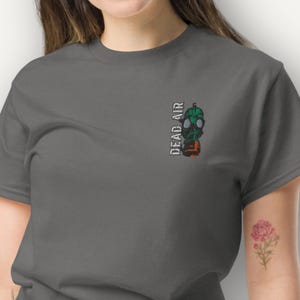 Puede incluir: Camiseta gris oscuro con un diseño de máscara de gas verde y detalles en naranja. El texto "DEAD AIR" está impreso verticalmente en el lado izquierdo. La camiseta es de un material suave.