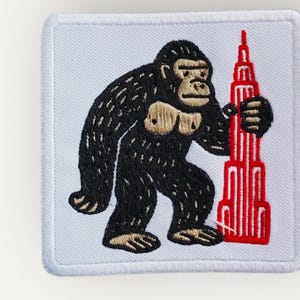 May include: A square embroidered patch with a white background and a white border. The design features a black gorilla with tan chest and hand details, standing next to a tall, red, multi-tiered skyscraper building illustration.