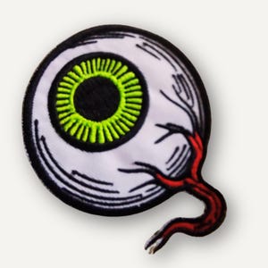 May include: Embroidered patch featuring a stylized eyeball. The eye has a black pupil, a bright green iris, and a white sclera. A red vein extends from the eye. The patch is outlined in black.