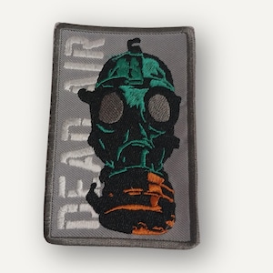 May include: Embroidered patch featuring a gas mask design in black, green, and orange on a gray rectangular background. The text "DEAD AIR" is vertically embroidered on the left side of the patch. The patch has a stitched border.
