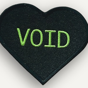 May include: Black heart-shaped patch with the word "VOID" embroidered in bright green. The patch has a textured surface and is likely designed to be sewn or ironed onto clothing or accessories.
