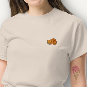 Op de afbeelding: Een beige t-shirt met een klein geborduurd ontwerp van een opgerolde oranje kat. De kat is gedetailleerd met zwarte en witte accenten. Het shirt heeft korte mouwen en een ronde hals.
