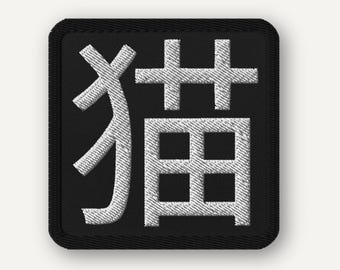 Peut inclure: Écusson brodé avec le caractère japonais 猫, signifiant "chat". Le design présente une couture blanche texturée sur un fond carré noir avec une bordure noire. Idéal pour personnaliser des vêtements ou des accessoires.