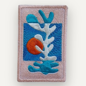 May include: A rectangular embroidered patch featuring a light pink border and a central blue square. Inside the square is an abstract white and teal branching shape alongside a solid orange circle, all rendered with dense, textured thread work.