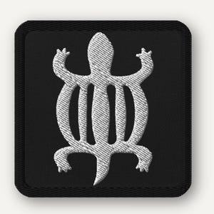 May include: Embroidered patch featuring a white Adinkra symbol, a stylized turtle, on a black square background. The symbol is intricately stitched, showcasing the texture of the thread. The patch has a black border.
