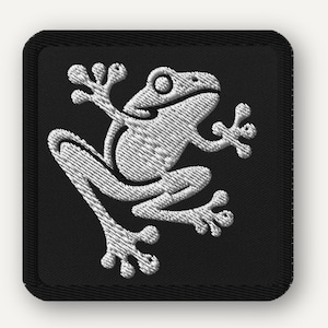 May include: Embroidered patch featuring a white frog design on a black square background. The frog is depicted in a stylized, detailed manner, with its limbs outstretched. The patch has a black border, suitable for sewing or ironing onto fabric.
