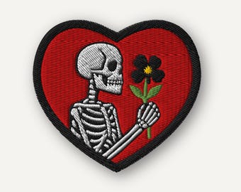Puede incluir: Parche bordado en forma de corazón con fondo rojo y borde negro. Presenta un esqueleto blanco sosteniendo una flor negra con hojas verdes. El esqueleto está de perfil.