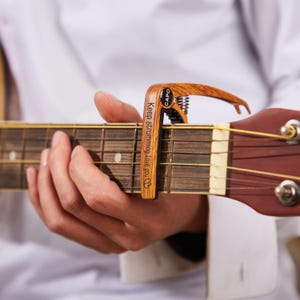 Op de afbeelding: Een houten gitaar capo met de tekst "Keep strumming, Love you" en het woord "CAPO" erop gedrukt. De capo is bevestigd aan de toets van een bruine akoestische gitaar. De capo heeft een veermechanisme.
