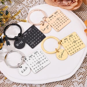 Puede incluir: Cuatro llaveros de plata y oro con dijes de calendario cuadrados y dijes redondos con texto personalizado. Los dijes de calendario presentan los meses de marzo, abril, mayo y julio de 2025 con un corazón que marca una fecha específica en cada calendario. Los dijes redondos tienen el texto "Love you", "W+M", "K&A" y "Sophia".