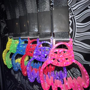 May include: Five colorful keychains with a glittery, eye-like design. Each keychain is made of a translucent plastic material and features a different color, including green, blue, purple, and pink.