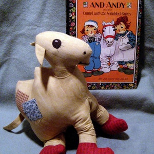 May include: A beige patchwork camel stuffed animal with red felt booties. The camel is sitting on a blue blanket next to a book titled "Raggedy Ann and Andy and the Camel with the Wrinkled Knees" by Johnny Gruelle.