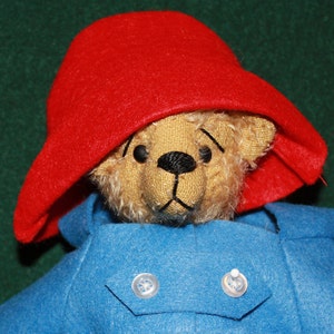 Può includere: Un orsacchiotto di peluche con un cappello rosso brillante e un cappotto blu con due bottoni bianchi. L'orso ha una faccia marrone con occhi neri e un naso e una bocca neri cuciti. Lo sfondo è verde scuro.