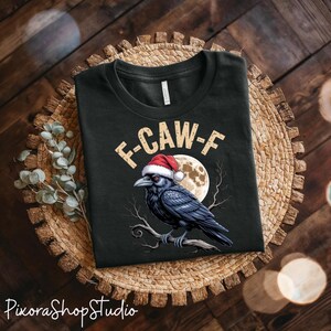 Puede incluir: Camiseta negra con un cuervo con un gorro de Papá Noel, posado en una rama, con una luna llena de fondo. El texto "F-CAW-F" está encima del cuervo. La camiseta se muestra sobre un salvamanteles tejido.