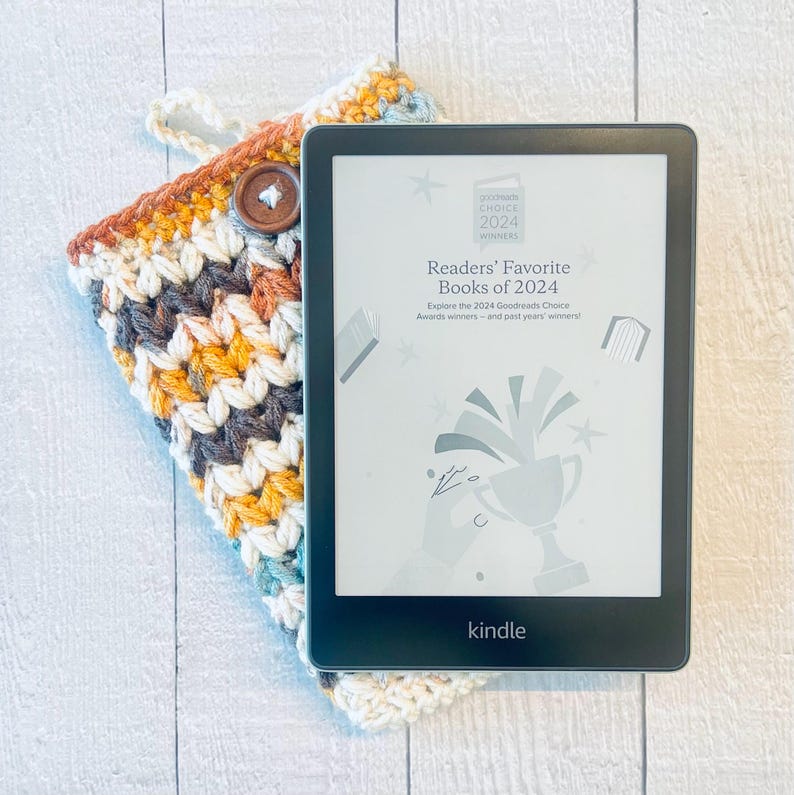 May include: A black and white Kindle e-reader with a white screen displaying the text "Readers' Favorite Books of 2024" and a graphic of a book being thrown into a trophy cup. The e-reader is resting on a crocheted book cover with a brown button.