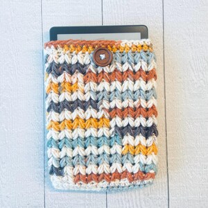 May include: A crocheted e-reader cover with a brown button closure. The cover is made with a multi-colored yarn in shades of white, gray, blue, orange, and yellow.