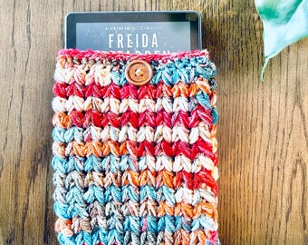 May include: A colorful, hand-crocheted e-reader sleeve with a wooden button closure. The sleeve features a vibrant mix of red, orange, blue, and cream-colored yarn. The e-reader displays the title "FREIDA" on the screen.