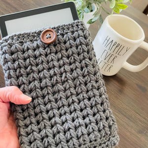 May include: Gray crocheted e-reader case with a wooden button. The case is made with a textured stitch pattern.
