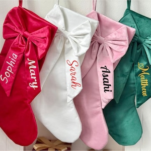 May include: Four velvet Christmas stockings in red, white, pink, and green, each with a large bow and a name embroidered on the ribbon. The names are Sophia, Mary, Sarah, Asahi, and Matthew.