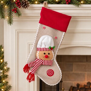 May include: A tan burlap Christmas stocking featuring a red cuff, white scalloped trim, and a plush gingerbread person appliqué wearing a white chef hat and a red and white striped scarf, accented with small red and white peppermint candy details.