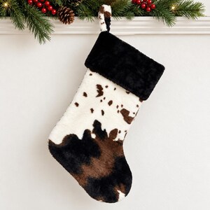 May include: A Christmas stocking made of plush faux fur featuring a white base with brown and black cow-print spots. The stocking has a solid black cuff at the top and a small loop for hanging, with a soft, textured appearance throughout.