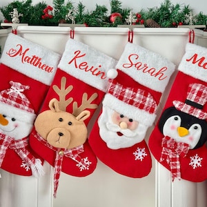 May include: Four red Christmas stockings with white fluffy tops hang above a fireplace. Each stocking has a name embroidered in red: Matthew, Kevin, Sarah, and Mary. The stockings are decorated with a snowman, reindeer, Santa, and penguin.