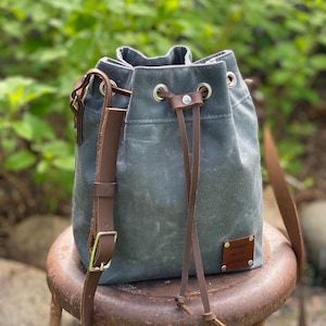 May include: A gray canvas bucket bag with brown leather straps and a drawstring closure. The bag has a small leather patch with the text "Nichole Designs" on it.