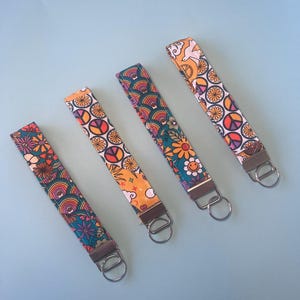 May include: Four patterned key fobs featuring rainbows, peace symbols, and floral designs. Each fob has a metal ring and a fabric strap. The key fobs are displayed against a light blue backdrop.