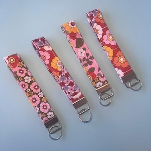 May include: Four floral fabric wrist keychains in shades of pink, red, and brown, each with a silver-coloured metal ring and hardware. The keychains feature a variety of flower patterns on a light blue background.