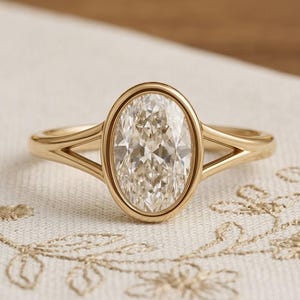 Op de afbeelding: Een gouden ring met een centrale, ovaal geslepen, heldere edelsteen in een gladde zetting. De ringband is gemaakt van gepolijst geelgoud en splitst zich in twee afzonderlijke armen waar deze aansluit op de zetting van de centrale steen.