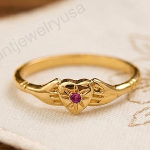 May include: A gold ring with a heart-shaped design and a small red gemstone in the center. The ring is on a white cloth with a floral pattern. The ring is a delicate piece of jewelry.