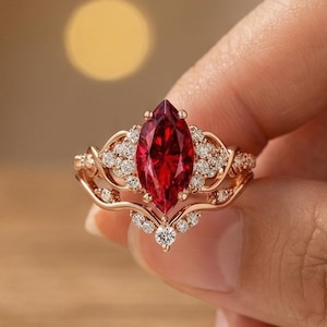 May include: A rose gold ring featuring a central marquise-cut deep red gemstone, flanked by smaller clear stones set along the band. The band has an intricate, curved design that wraps around the central stone, creating a layered appearance.