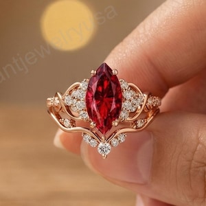 May include: A rose gold ring featuring a marquise-cut, deep red gemstone as the centerpiece. The ring is adorned with small, clear accent stones and intricate filigree detailing. The ring is held against a soft, neutral background.