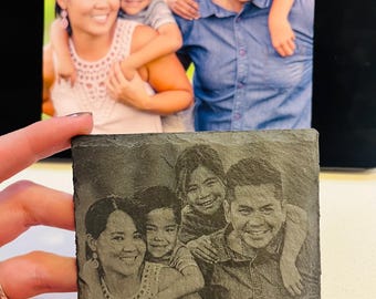 May include: A family portrait is displayed on a slate coaster. The coaster features a laser-etched image of the family, mirroring the color photograph behind it. The family consists of two adults and two children, all smiling.