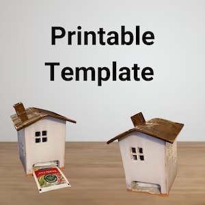 May include: Printable template for a ceramic tea bag holder shaped like a house. The house is white with a brown roof and a small window. The tea bag holder is sitting on a wooden surface. The text "Printable Template" is written in black letters on a white background.
