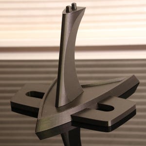 May include: A black 3D printed stand with a triangular base and a curved, vertical support. The stand is designed to hold a small object, such as a figurine or a model.