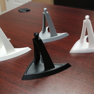 Puede incluir: Cuatro expositores impresos en 3D en blanco, gris y negro. Cada soporte tiene una base triangular y un soporte vertical con una parte superior redondeada. Los soportes están diseñados para sostener un artículo en posición vertical.