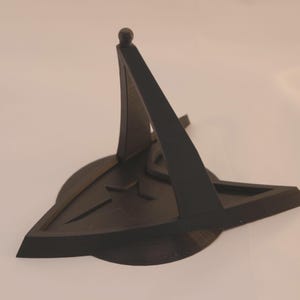 Puede incluir: Un soporte de exhibición de plástico negro con forma del emblema de la Flota Estelar de Star Trek. El soporte tiene una sección central elevada para exhibir una figura u otro objeto.