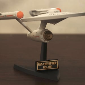 May include: A model of the USS Enterprise NCC-1701 starship featuring a white saucer section, two long engine nacelles with orange caps, and a central deflector dish. The model is mounted on a black stand with a plaque reading USS ENTERPRISE NCC-1701.