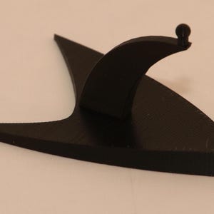 Op de afbeelding: Een zwarte plastic standaard in de vorm van het Star Trek Starship Enterprise-insigne. De standaard heeft een verhoogd, gebogen gedeelte aan de achterkant om een klein object te tonen.