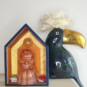 Può includere: Una piccola casetta di legno con bordi blu e dettagli dorati. All'interno della casa c'è una statuetta di una ragazza che indossa un vestito. La casa è appoggiata su un tavolo accanto a una statuetta di tucano nero e verde con un becco dorato.