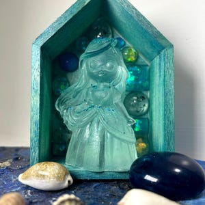 Può includere: Una scatola di legno blu-verde a forma di casa con una statuetta di una principessa in vetro trasparente all'interno. La scatola è piena di perline di vetro blu, verdi e iridescenti. La principessa indossa una corona e un lungo abito svolazzante. La scatola è appoggiata su una superficie blu e bianca con conchiglie.