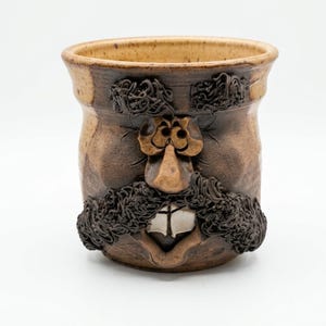 May include: A handmade ceramic mug with a face design. The mug is brown with a tan interior and features a sculpted face with a mustache, beard, and eyebrows. The mouth is open, revealing two teeth.