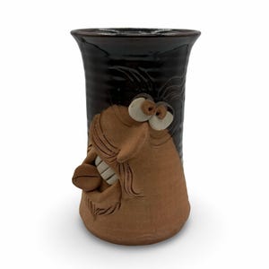 May include: A handcrafted ceramic vase with a comical face design. The vase has a brown face with exaggerated features, including large eyes, a prominent nose, and a smiling mouth. The upper part of the vase is dark brown.