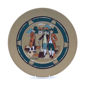 May include: A decorative plate featuring a tan border and a central image of three men in historical attire. The image is framed by a teal and black geometric design. The plate has the text "Ye Olden Times".