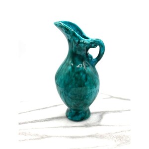 Peut inclure: Un petit pichet en céramique de couleur teal, avec une anse et un bec verseur incurvé. Le pichet a une finition brillante et un corps bulbeux, reposant sur une petite base. La poignée est en forme de boucle.