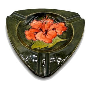 May include: A triangular, green ceramic ashtray featuring an orange hibiscus flower design. The flower is detailed with silver outlines and green leaves. The ashtray has three cigarette rests.