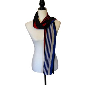 May include: A long scarf featuring a colour-blocked design with a black, red, and white striped pattern on one end and a blue, white, and black vertical striped pattern on the other, draped around a white mannequin torso for display purposes.