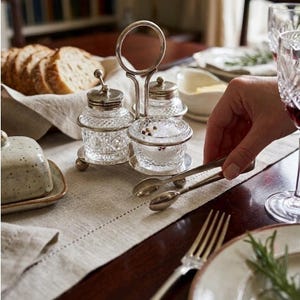 Peut inclure: Un ensemble sel et poivre argenté avec un support décoratif. L'ensemble comprend deux pots à couvercle et un petit bol ouvert, tous en verre taillé transparent. La table comprend également un beurrier, du pain et un verre à vin.