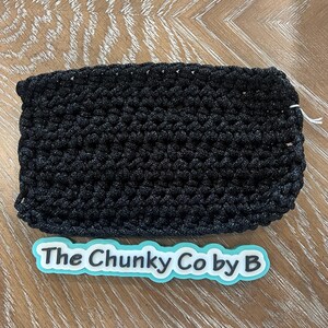 May include: A black, rectangular, crocheted item. The item is made of a thick, textured yarn. The text "The Chunky Co by B" is on a light blue and white sticker below the item. The item is on a wooden surface.