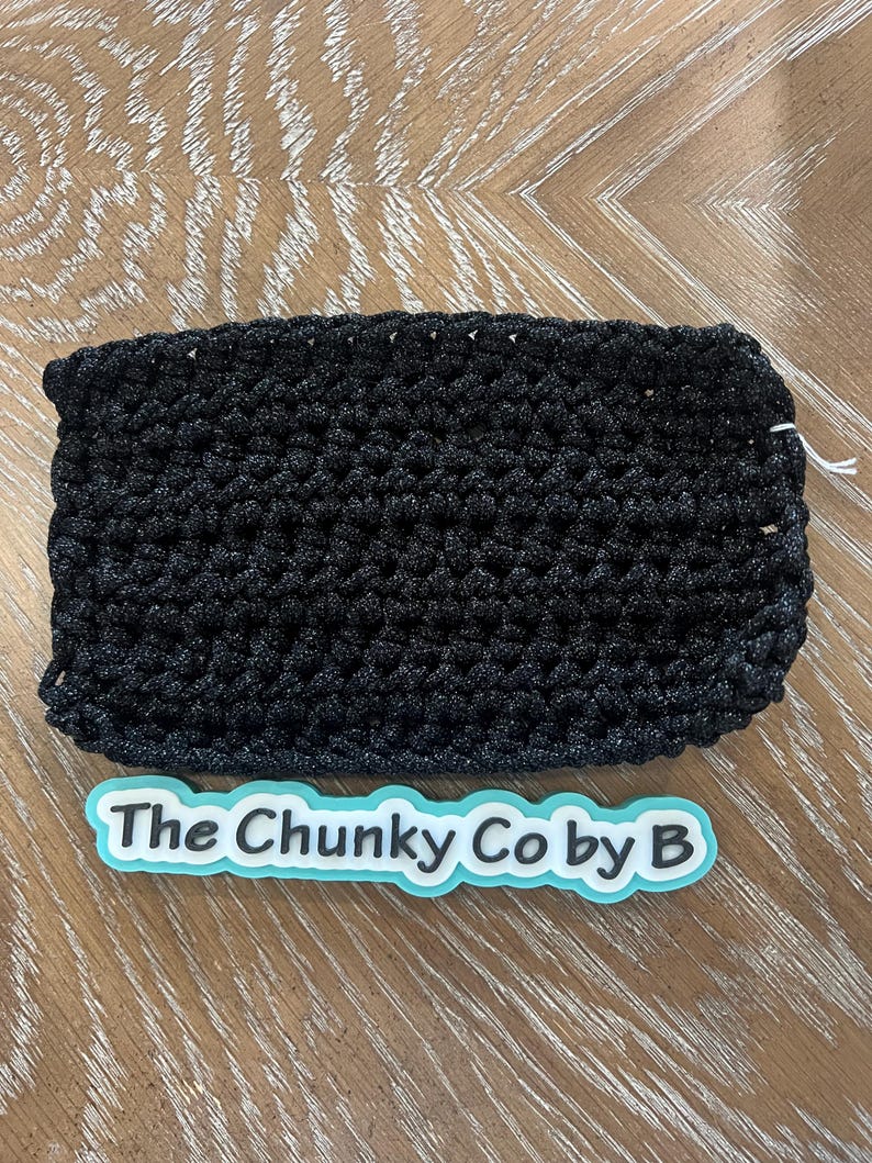 May include: A black, rectangular, crocheted item. The item is made of a thick, textured yarn. The text "The Chunky Co by B" is on a light blue and white sticker below the item. The item is on a wooden surface.
